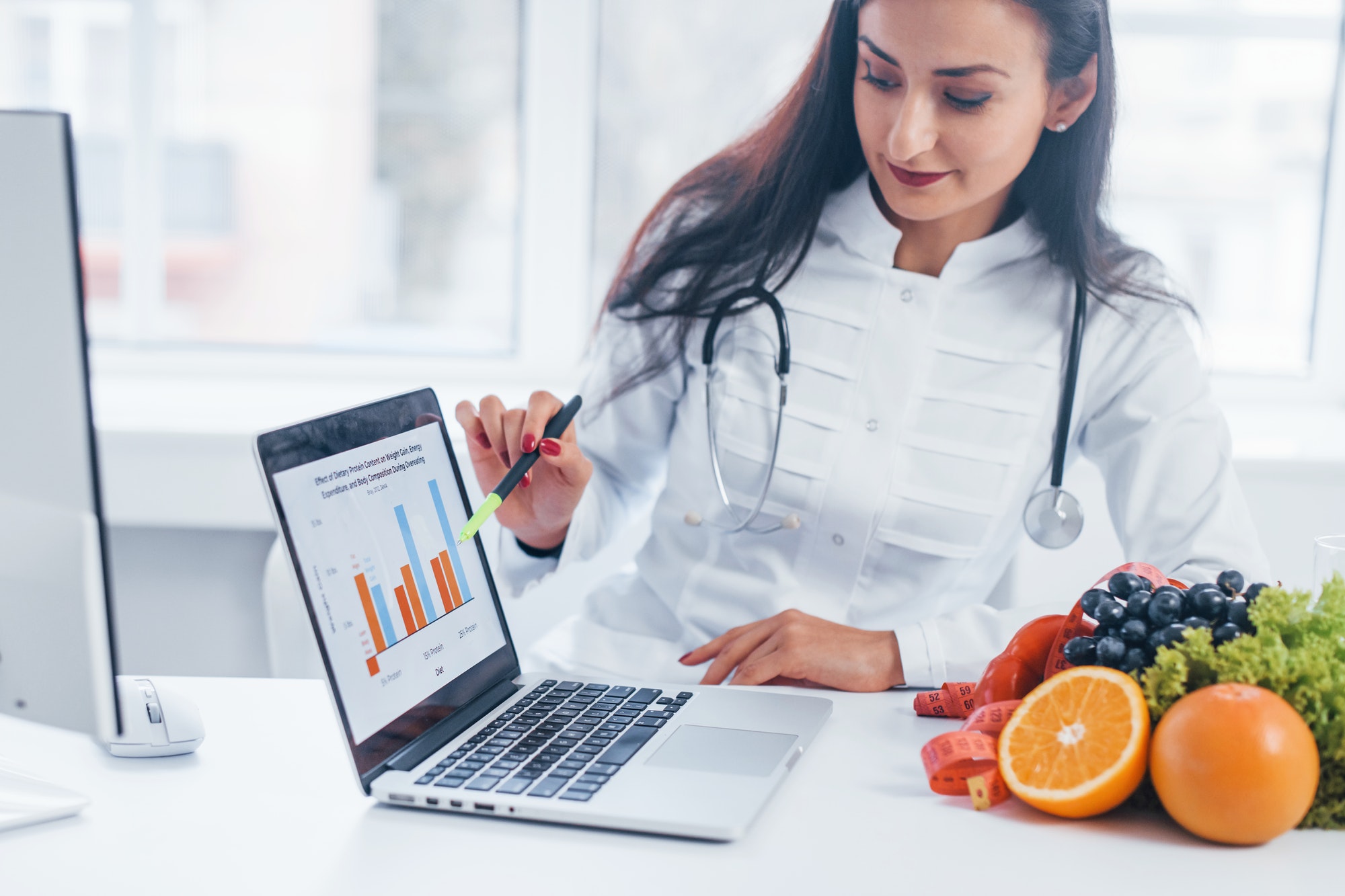 Female nutritionist in white coat sitting indoors in the office at workplace with laptop Female nutritionist in white coat sitting indoors in the office at workplace with laptop