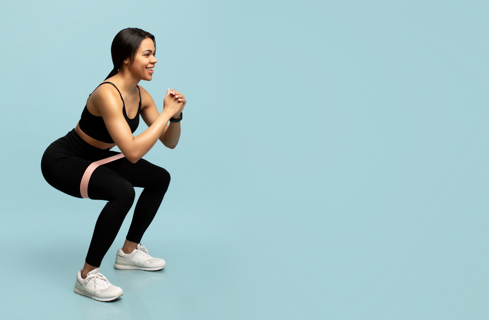 Training for glute muscles. Sporty black woman doing deep squat exercises with elastic bands, blue
